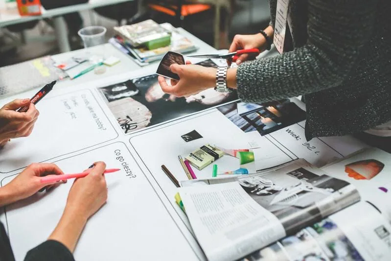 Kreative Corporate Design Entwicklung bei Sofinity – Zwei Mitarbeiter gestalten mit Schere, Büchern und Designmaterialien am Tisch in Landsberg am Lech